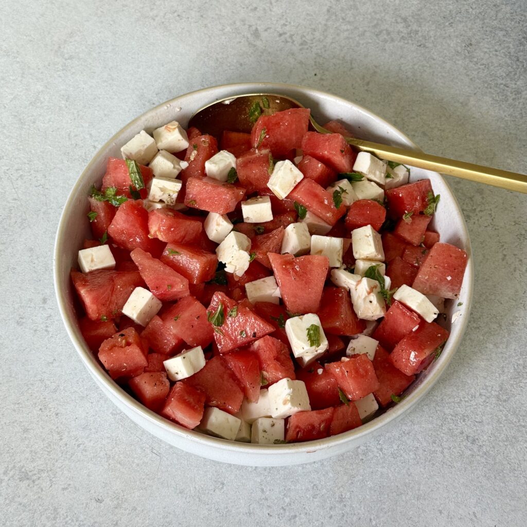 Der Wassermelonen-Feta Salat ist fertig in einer weißen Schüssel angerichtet. Ein goldener Löffel ist dabei.